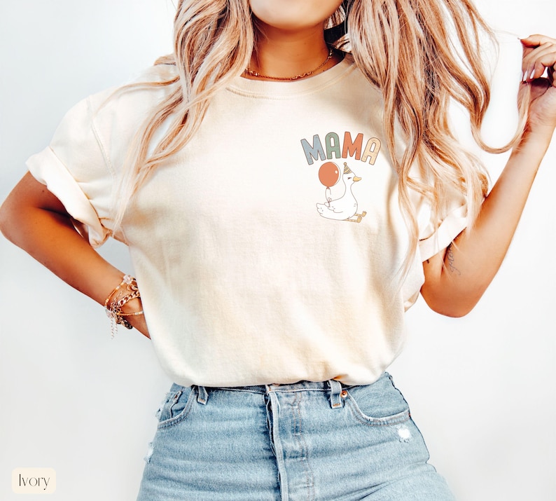 Puede incluir: Camiseta de color marfil con la palabra "MAMA" en letras pastel y un dibujo de un ganso sosteniendo un globo. El ganso lleva un gorro de fiesta. La camiseta se combina con vaqueros azules.