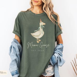 May include: A green t-shirt with a white goose wearing a party hat and the text "Mama Goose Est 2024".