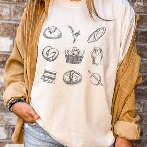 Peut inclure: T-shirt &agrave; manches longues de couleur ivoire avec un imprim&eacute; graphique de divers articles de boulangerie et de pain, notamment un pain, un bretzel, un panier de pain, un pot de p&acirc;te, un rouleau &agrave; p&acirc;tisserie et des &eacute;pis de bl&eacute;.