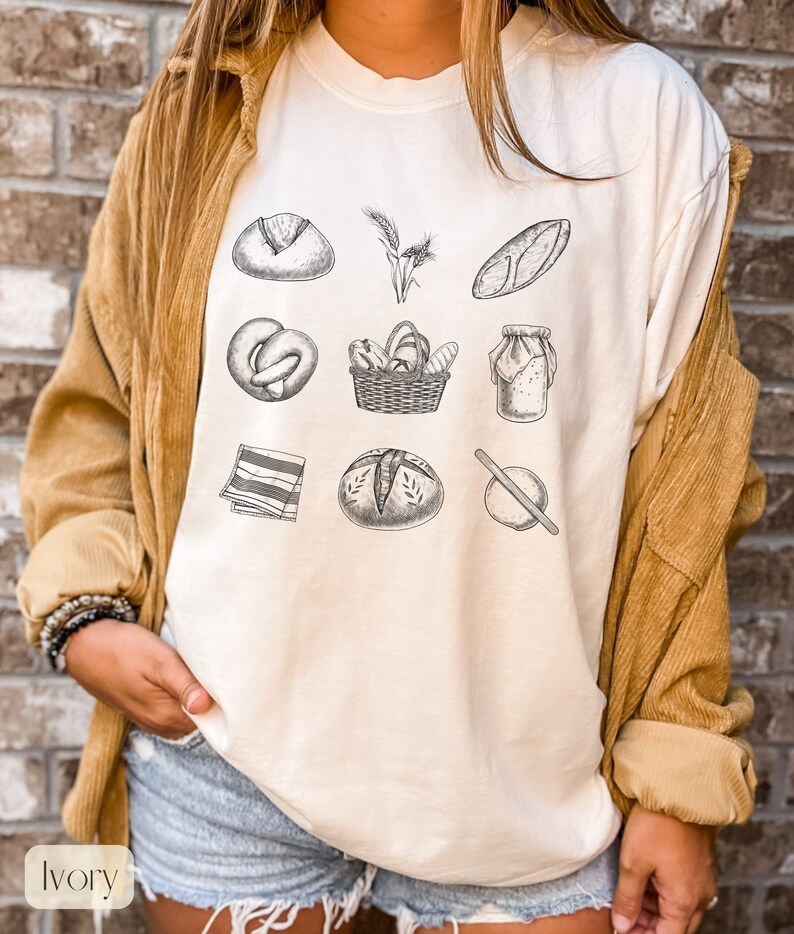 Peut inclure: T-shirt &agrave; manches longues de couleur ivoire avec un imprim&eacute; graphique de divers articles de boulangerie et de pain, notamment un pain, un bretzel, un panier de pain, un pot de p&acirc;te, un rouleau &agrave; p&acirc;tisserie et des &eacute;pis de bl&eacute;.