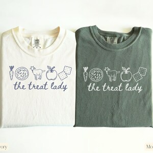 Puede incluir: Dos camisetas, marfil y verde musgo, con cuello redondo y las palabras "the treat lady" en letra cursiva. Las camisetas presentan un dibujo lineal de una zanahoria, un donut, una cabra, una manzana y galletas.