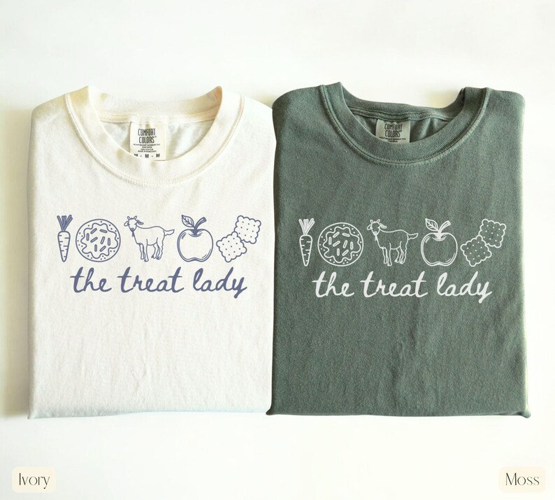 Puede incluir: Dos camisetas, marfil y verde musgo, con cuello redondo y las palabras "the treat lady" en letra cursiva. Las camisetas presentan un dibujo lineal de una zanahoria, un donut, una cabra, una manzana y galletas.