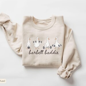 K&ouml;nnte beinhalten: Sandfarbenes Sweatshirt mit einer Grafik von vier Cartoon-G&auml;nsen, die Gewichte heben. Der Text "barbell baddie" ist darunter in einer handschriftlichen Schriftart aufgedruckt. Das Sweatshirt hat einen Rundhalsausschnitt und lange &Auml;rmel.