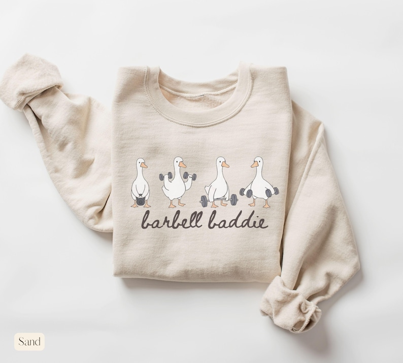 K&ouml;nnte beinhalten: Sandfarbenes Sweatshirt mit einer Grafik von vier Cartoon-G&auml;nsen, die Gewichte heben. Der Text "barbell baddie" ist darunter in einer handschriftlichen Schriftart aufgedruckt. Das Sweatshirt hat einen Rundhalsausschnitt und lange &Auml;rmel.