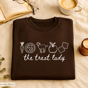 Puede incluir: Sudadera marr&oacute;n chocolate con un gr&aacute;fico blanco de una zanahoria, una rosquilla, una cabra, una manzana y galletas, con el texto "the treat lady". La sudadera est&aacute; doblada sobre una superficie beige, con una vela encendida, un libro abierto y caf&eacute;.