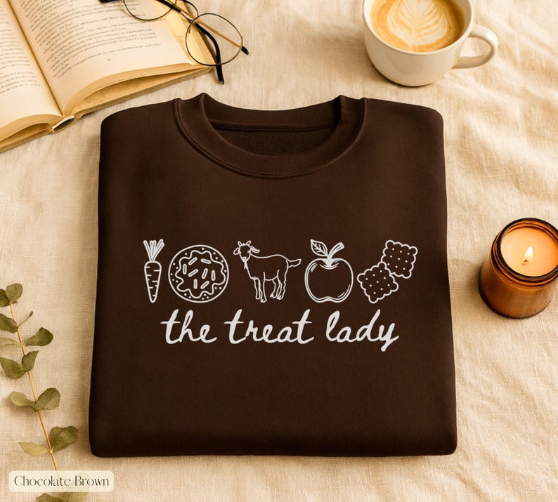 Puede incluir: Sudadera marr&oacute;n chocolate con un gr&aacute;fico blanco de una zanahoria, una rosquilla, una cabra, una manzana y galletas, con el texto "the treat lady". La sudadera est&aacute; doblada sobre una superficie beige, con una vela encendida, un libro abierto y caf&eacute;.