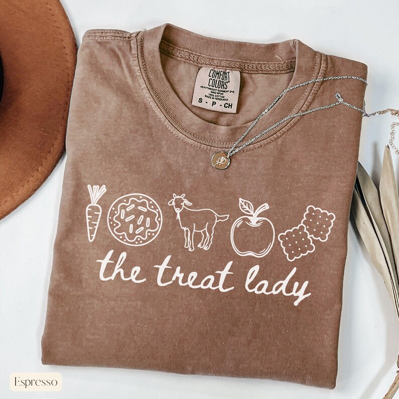 Puede incluir: Camiseta color espresso con el texto "the treat lady" e ilustraciones blancas de una zanahoria, una rosquilla, una cabra, una manzana y galletas saladas. Un collar plateado con un colgante est&aacute; sobre la camiseta.