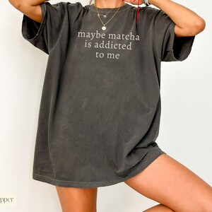 Puede incluir: Camiseta gris oscuro de gran tama&ntilde;o con el texto blanco "maybe matcha is addicted to me". La camiseta tiene mangas cortas y un corte holgado, perfecta para un look casual. La tela parece suave y ligeramente descolorida.