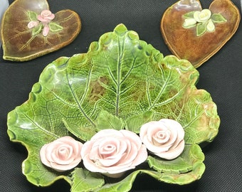 May include: Three ceramic dishes shaped like leaves with raised floral details. The center dish is green with three pink roses, the other two are brown with pink flowers.