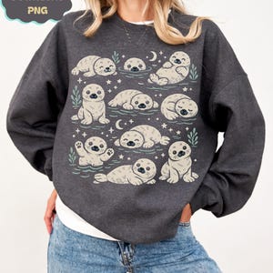 Op de afbeelding: Donkergrijze sweatshirt met een herhaald patroon van cartoon zeehonden in verschillende poses. De zeehonden zijn lichtbeige met zwarte ogen en snorharen. Het ontwerp bevat sterren, halve manen en takjes groen. De tekst "DOWNLOAD PNG" staat linksboven.