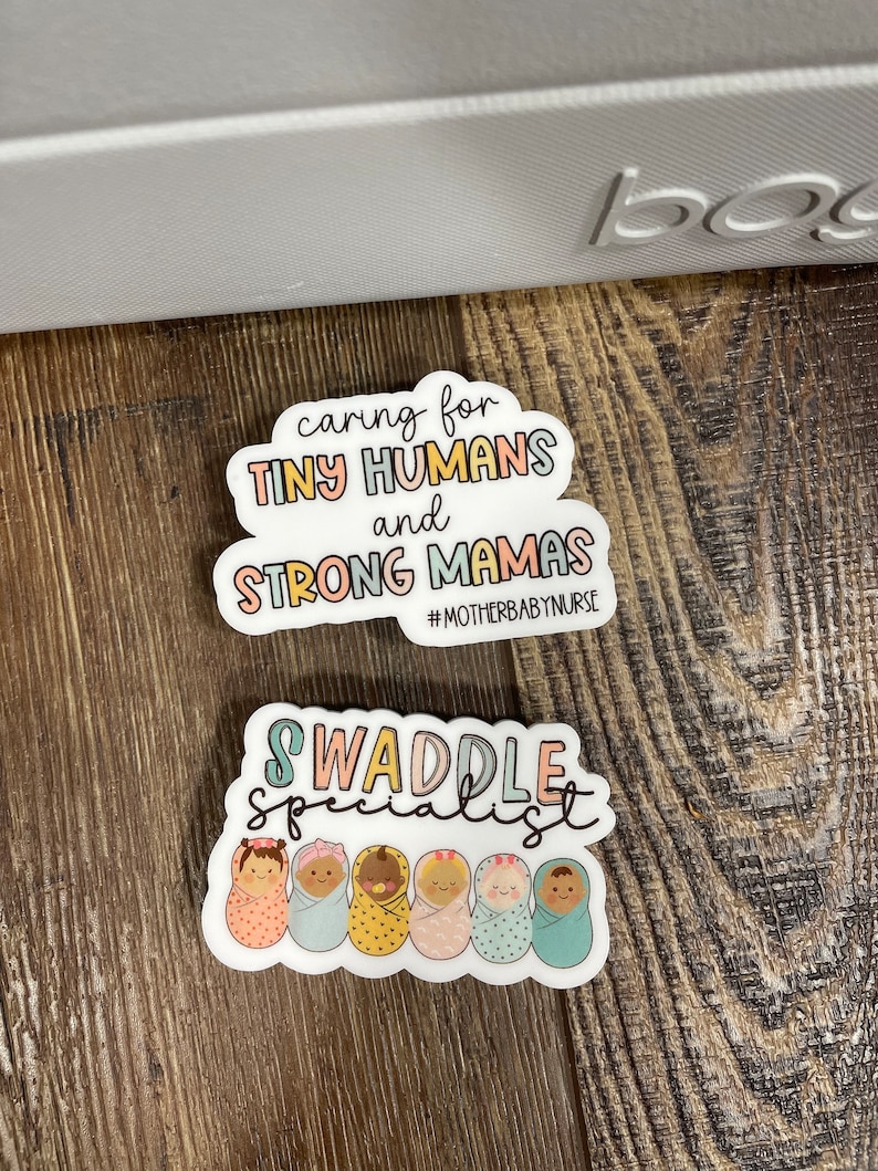 May include: Two stickers with a baby theme. The first sticker is white with a pink, blue, and yellow cloud shape. The text on the sticker reads "caring for TINY HUMANS and STRONG MAMAS #MOTHERBABY NURSE". The second sticker is white with a pink, blue, and yellow swirly design. The text on the sticker reads "SWADDLE SPECIALIST" and features a row of six cartoon babies in swaddles.