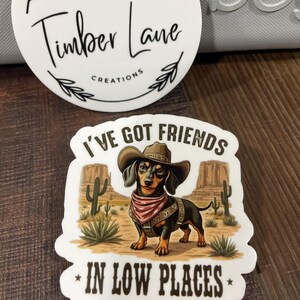 May include: A white sticker with a brown dachshund wearing a cowboy hat and bandana. The text on the sticker reads "I've got friends in low places".
