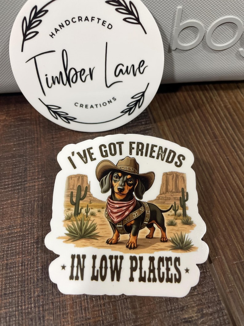 May include: A white sticker with a brown dachshund wearing a cowboy hat and bandana. The text on the sticker reads "I've got friends in low places".