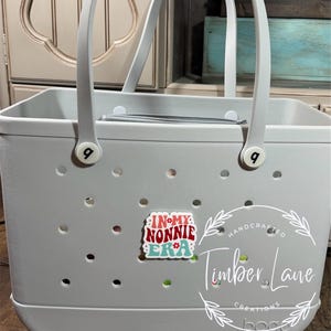 May include: A light gray tote bag with white handles and the text "IN MY NONNIE ERA" on a decorative charm. The bag has a perforated design and the brand name "bogg" is visible.