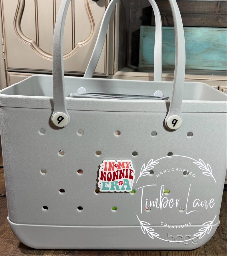 May include: A light gray tote bag with white handles and the text "IN MY NONNIE ERA" on a decorative charm. The bag has a perforated design and the brand name "bogg" is visible.