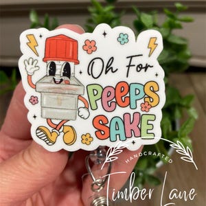 May include: A white badge reel featuring a cartoon lightbulb character with the text "Oh For Peeps Sake". The lightbulb has a red top and yellow shoes, with flowers and lightning bolts. The badge reel is from Timber Lane Creations.