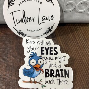 May include: A white sticker with a blue bird cartoon with a tongue sticking out. The text on the sticker reads "Keep rolling your EYES you might find a BRAIN back there."