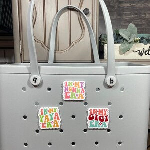 May include: A gray tote bag with the word "bogg" printed on the bottom. The bag has three colorful stickers on the front that say "In My Nonna Era", "In My Yaya Era", and "In My Gigi Era".