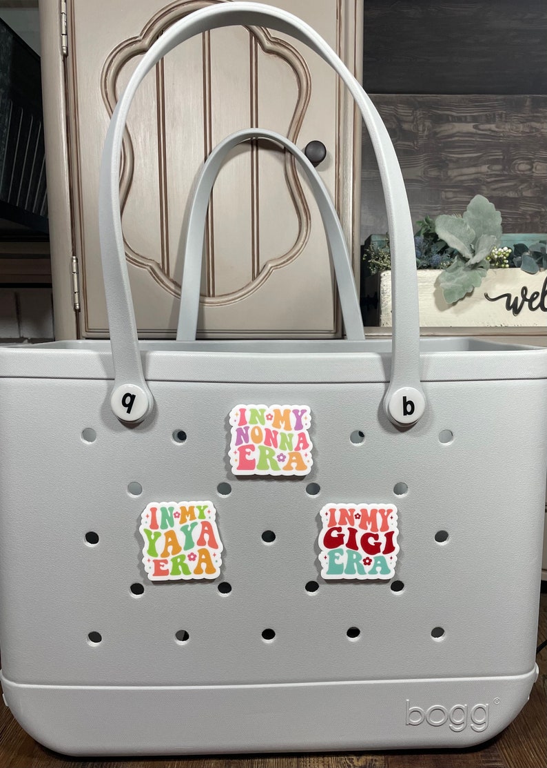 May include: A gray tote bag with the word "bogg" printed on the bottom. The bag has three colorful stickers on the front that say "In My Nonna Era", "In My Yaya Era", and "In My Gigi Era".
