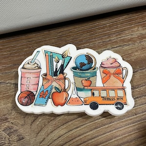 May include: A sticker featuring a school bus, a globe, a cup of coffee, a cup of hot chocolate, a pencil, a paintbrush, a book, an apple, and a beaker. The sticker has a white background and the text "School Bus" is printed on the bus.