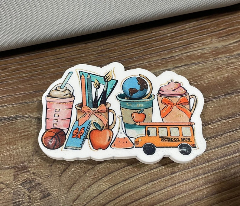 May include: A sticker featuring a school bus, a globe, a cup of coffee, a cup of hot chocolate, a pencil, a paintbrush, a book, an apple, and a beaker. The sticker has a white background and the text "School Bus" is printed on the bus.