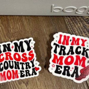 May include: Two white stickers with red and black text. The first sticker says "In My Cross Country Mom Era" with a red heart. The second sticker says "In My Track Mom Era" with a red winged foot.