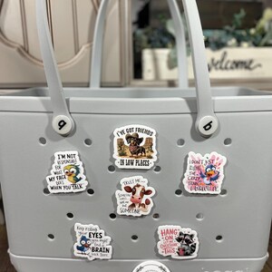 May include: A gray tote bag with a variety of colorful stickers featuring animals and humorous sayings. The stickers include a cow, a raccoon, a bird, and a dachshund. The bag has a white label with the word "bogg" printed on it.