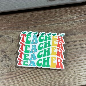 May include: A sticker with the word "TEACHER" repeated four times in a stack. Each word is in a different color: red, yellow, green, and blue. The letters are stylized and have a retro look. Each word has a small green leaf on top.