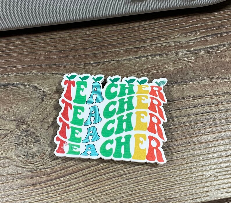 May include: A sticker with the word "TEACHER" repeated four times in a stack. Each word is in a different color: red, yellow, green, and blue. The letters are stylized and have a retro look. Each word has a small green leaf on top.