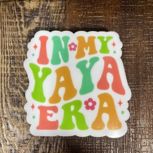 May include: A white sticker with the text "In My Yaya Era" in colorful, retro-style lettering. The letters are in shades of orange, green, and pink.