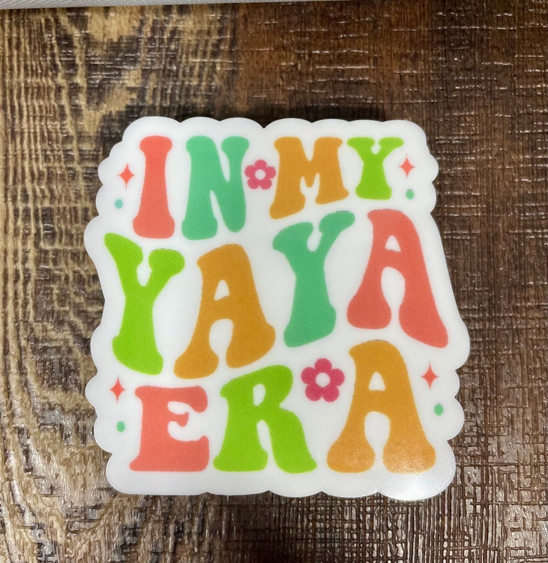 May include: A white sticker with the text "In My Yaya Era" in colorful, retro-style lettering. The letters are in shades of orange, green, and pink.