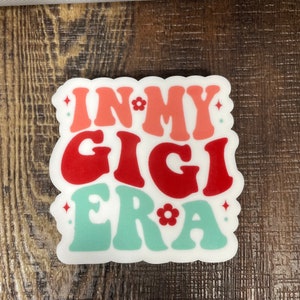 May include: White sticker with the text "In My Gigi Era" in a retro font. The words "In" and "Era" are in pink, "My" is in white, and "Gigi" is in red. There are small white flowers and stars around the text.