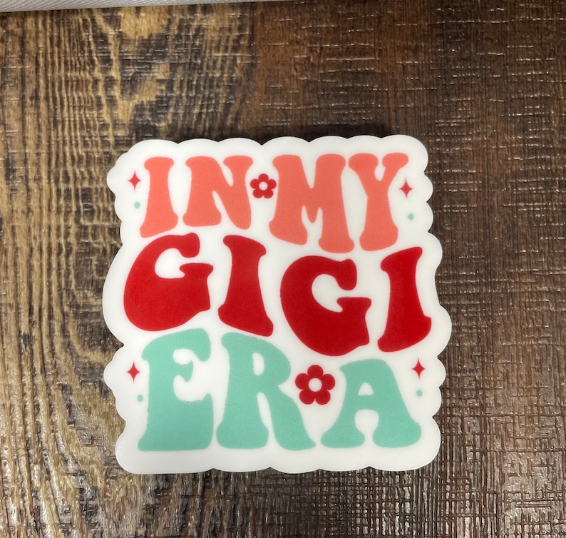 May include: White sticker with the text "In My Gigi Era" in a retro font. The words "In" and "Era" are in pink, "My" is in white, and "Gigi" is in red. There are small white flowers and stars around the text.