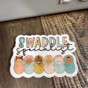 May include: A white sticker with the text "Swaddle Specialist" in rainbow colours. Six cartoon babies are swaddled in different coloured blankets with patterns. The babies have different skin tones and hair colours.