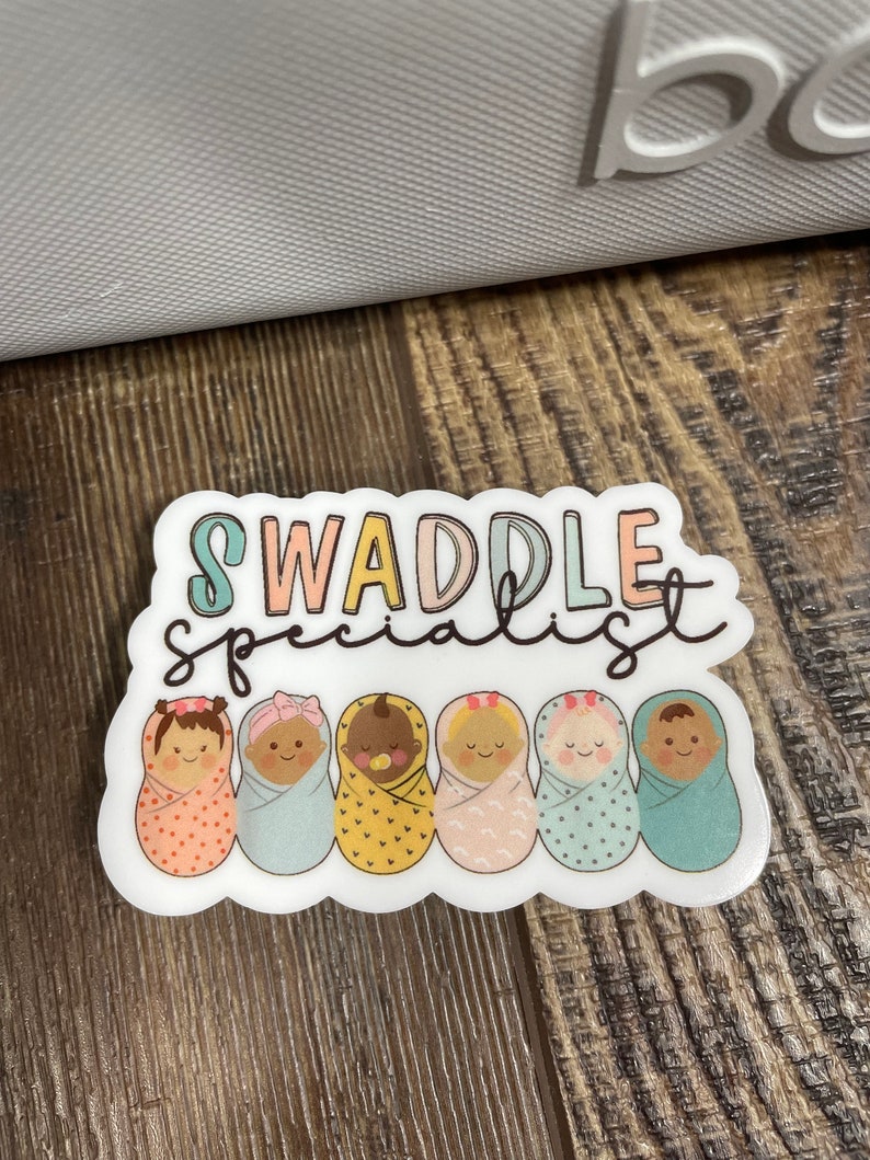 May include: A white sticker with the text "Swaddle Specialist" in rainbow colours. Six cartoon babies are swaddled in different coloured blankets with patterns. The babies have different skin tones and hair colours.