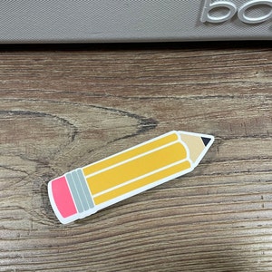 May include: A yellow pencil sticker with a pink eraser and a black tip. The sticker is on a brown wooden surface.