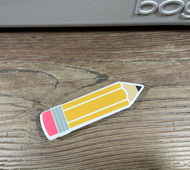 May include: A yellow pencil sticker with a pink eraser and a black tip. The sticker is on a brown wooden surface.