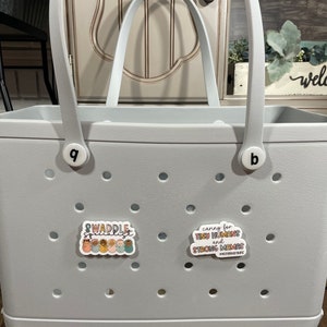 May include: A grey tote bag with the word "BOGG" printed on the bottom. The bag has two handles and a white sticker that says "caring for tiny humans and strong mamas #motherhoodmagic". There is another sticker on the bag that says "Swaddle Specialist" with a graphic of several babies in swaddles.