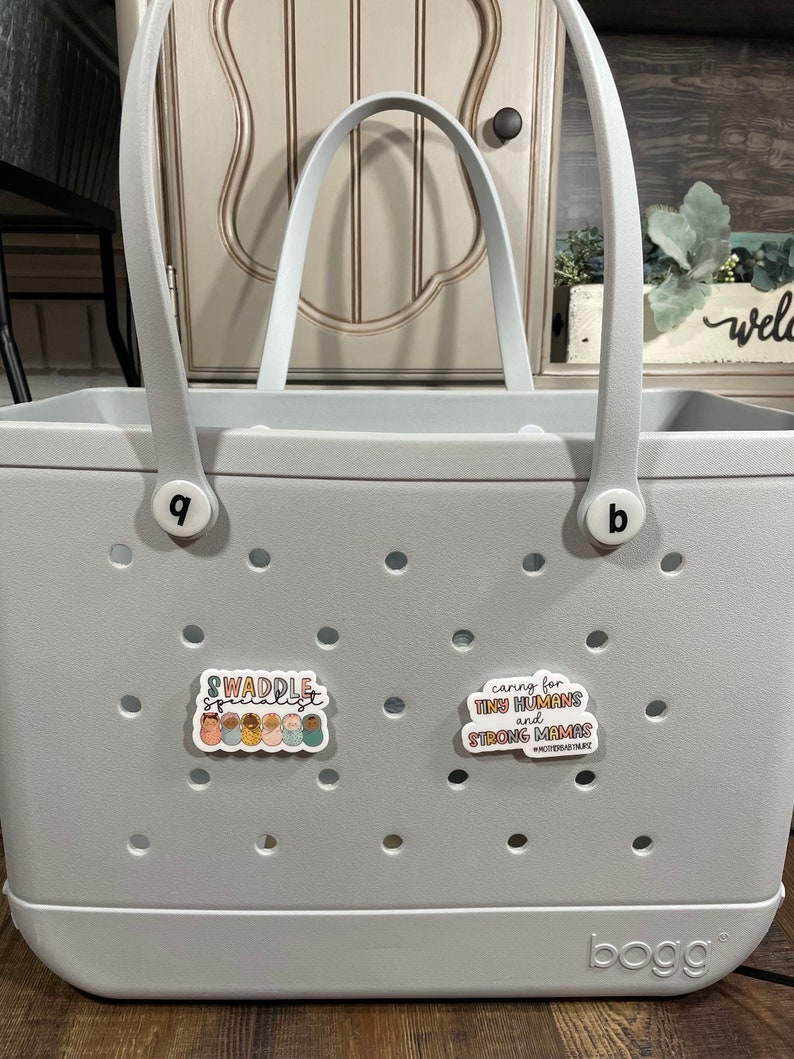 May include: A grey tote bag with the word "BOGG" printed on the bottom. The bag has two handles and a white sticker that says "caring for tiny humans and strong mamas #motherhoodmagic". There is another sticker on the bag that says "Swaddle Specialist" with a graphic of several babies in swaddles.