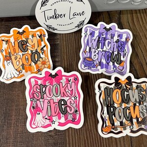 May include: Four Halloween-themed stickers with phrases like "Hey Boo" and "Spooky Vibes." Each sticker features a unique design with bats, ghosts, and other spooky elements. The stickers have a white border and are set against a wooden surface.