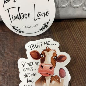 May include: White sticker with a brown and white cow illustration. The cow has a pink nose and is looking at the viewer. The text on the sticker reads "Trust me... Someone cares... Not me but Someone".