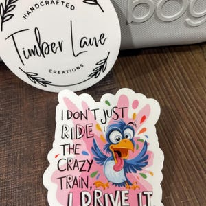 May include: A white sticker with a pink background and a cartoon blue bird with a red tongue. The text on the sticker reads "I don't just ride the crazy train, I drive it."