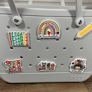 May include: A gray tote bag with a variety of colorful stickers. The stickers feature a rainbow with pencils, a coffee cup, a teacher with a stack of books, and the text "#Teacherlife".