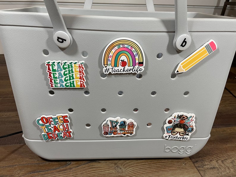 May include: A gray tote bag with a variety of colorful stickers. The stickers feature a rainbow with pencils, a coffee cup, a teacher with a stack of books, and the text "#Teacherlife".