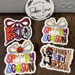 May include: A collection of Halloween-themed stickers. Designs include the word "BOO" with a bow, "Spooky Season" with ghosts and a pumpkin, and a raccoon with candy corn. The stickers are white with colorful accents.