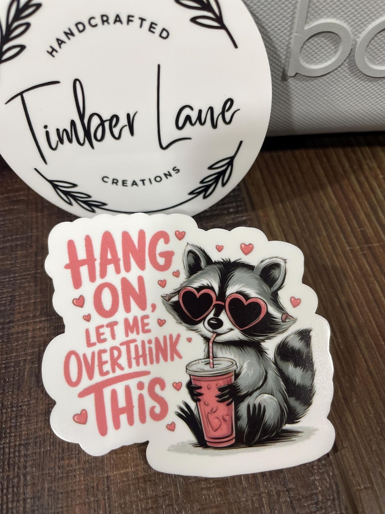 May include: White sticker with a cartoon raccoon wearing heart-shaped sunglasses and drinking from a pink cup. The text on the sticker reads "Hang on, let me overthink this".