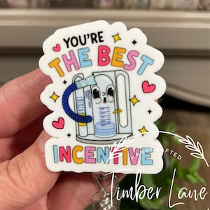 May include: A white badge reel with the text "YOU'RE THE BEST INCENTIVE" in various colours. The badge features a cartoon image of a medical device and decorative hearts and stars. The badge is held by a person.