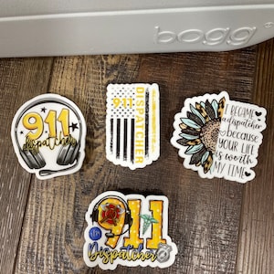 May include: Four 911 dispatcher themed stickers. One sticker features a headset with the number 911 and the word "dispatcher" written in a yellow banner. Another sticker features a yellow and black American flag with the number 911 and the word "dispatcher" written vertically on the right side. A third sticker features a sunflower with a leopard print design and the text "I became a dispatcher because your life is worth my time". The final sticker features a yellow 911 with a firefighter helmet, a medical symbol, and a police badge. The text "Dispatcher" is written in a yellow banner below the number.