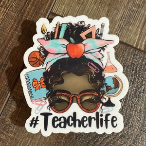 May include: A white sticker with a cartoon illustration of a Black woman with a red apple, a blue and pink bow, and a variety of school supplies in her hair. The text "#Teacherlife" is below the illustration.