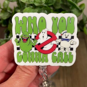 May include: A white badge reel with the text "WHO YOU GONNA CALL" in green, featuring cartoon characters from the Ghostbusters franchise. The reel includes a green Slimer, a ghost, and the Stay Puft Marshmallow Man.